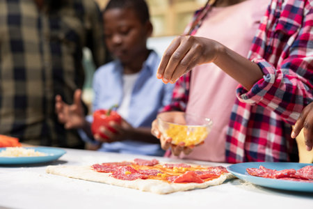 Close up of family fun in the kitchen as everyone is layering cheese and mozzarella on the dough, preparing homemade pizza together. Warm domestic scene captures joy and delicious homemade food.の写真素材