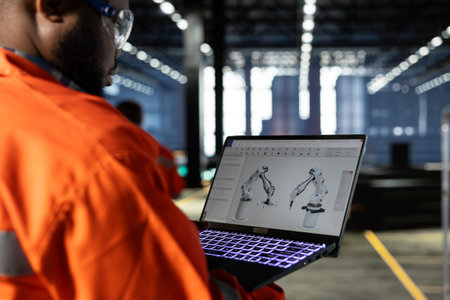 Black employee using automation software connected to robot machinery via laptop, surrounded by metal equipment and fabrication tools. Industrial environment highlights engineering.の写真素材
