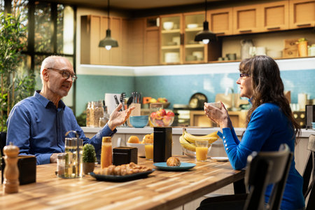 Elderly couple enjoys natural morning routine with healthy homemade breakfast, eating pastry and drinking coffee. Husband and wife shares casual conversation for candid moment.の写真素材