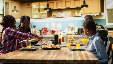 Cheerful black family enjoying breakfast together in a bright cozy kitchen. Parents serve toast and...