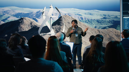 Black female speaker does wind power presentation at business forum with wind turbine machine and rotation. Addressing listeners during public speaking event focused on alternative energy.の写真素材