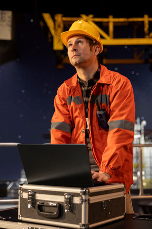 Professional male engineer in hard hat performing analysis on laptop from offshore platform,...