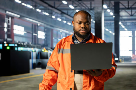 Black industrial personnel overseeing workshop fabrication equipment on laptop, emphasizing heavy industry safety and advanced development with automation in the manufacturing process.の写真素材