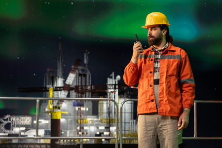 Drilling ship employee coordinates remote team tasks using portable radio, aurora borealis background. Specialist speaks into walkie talkie, managing offshore platform logistics under northern lightsの写真素材