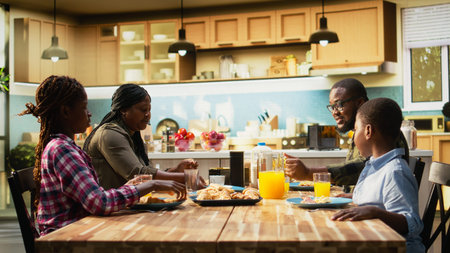 African American family sharing eggs and pastry for breakfast at home, parents serve omelettes, toast and fruit. Cozy kitchen moment captures warmth, relaxation and joyful togetherness. Camera A.の写真素材