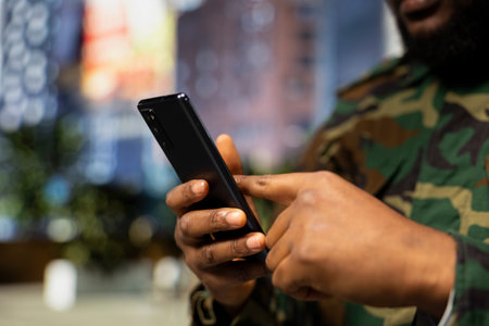 Close up of black male service person in full gear opens mobile phone map, walking through a modern downtown area in uniform. Soldier admiring skyscrapers and busy city street life.の写真素材