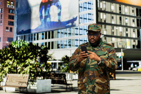 Adult male soldier in uniform navigates mobile screen on downtown street, showing disciplined confidence and respectful presence as he checks online page near urban buildings.の写真素材