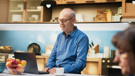 Mature pensioner enjoys happy video conference with family on his laptop. Senior man relaxing with coffee and pastry, showing a positive online communication during retired life. Camera B.の写真素材