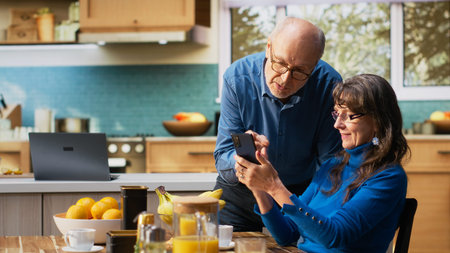 Senior woman and her husband laughing and checking smartphone at the breakfast table to enjoy a cozy meal with toast and coffee. Chatting and reflecting warm retirement lifestyle. Camera B.の写真素材