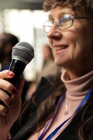 Close up of old female attendee asking questions on microphone and interacting during discussion, engaging in debate at international summit. Business conference with presentation on stage.の写真素材