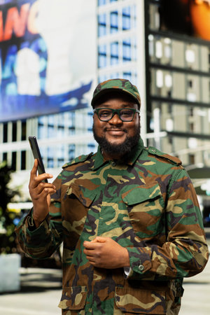 Black adult male in military uniform chatting on phone call with his officer, walking around city center in patrol. Male soldier stands confidently near skyscrapers downtown, metropolis.の写真素材
