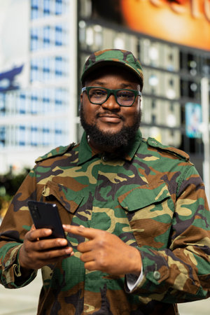 African american soldier in uniform using a mobile phone on a city street, scrolling a web page as he maintains disciplined posture and strong commitment to duty in downtown area.の写真素材