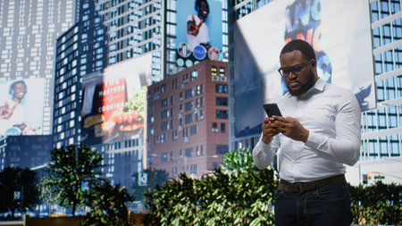Adult male scrolling on his mobile phone walking along city sidewalk in bright day sunlight,...