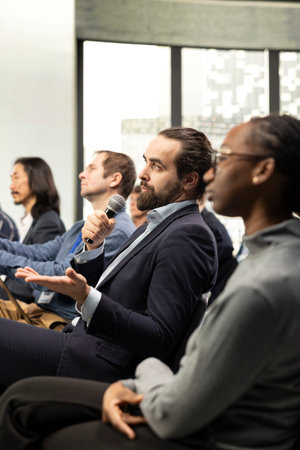 Male CEO in attendance asking questions during conference discussion, engaging in professional debate and listening to speech. Group of white collar workers attend international summit.の写真素材