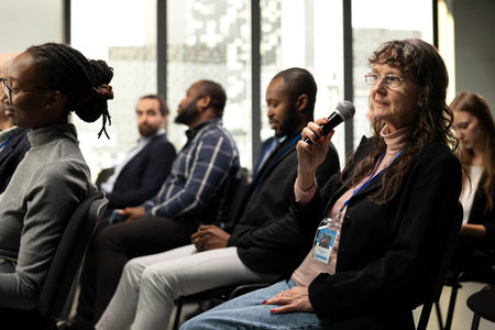 Senior woman participant in the audience asking questions during conference, interacting in...
