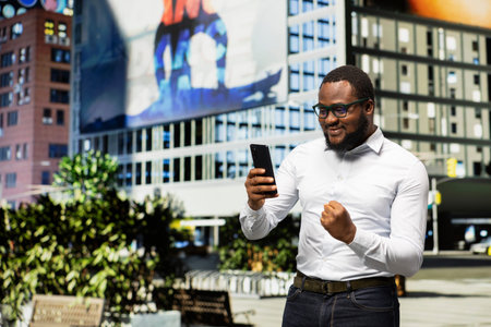 Black adult being happy and pleased with good news on phone screen, walking on a street downtown and...