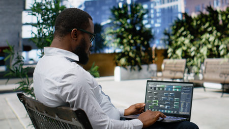 African american male engaged in reading data analysis on laptop and sitting on a sidewalk bench,...