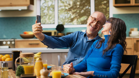 Elderly couple taking pictures on mobile phone during casual breakfast at the kitchen table with...