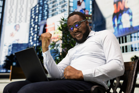 Black male adult being excited and cheering for his accomplishment outdoor on a bench, reading good...