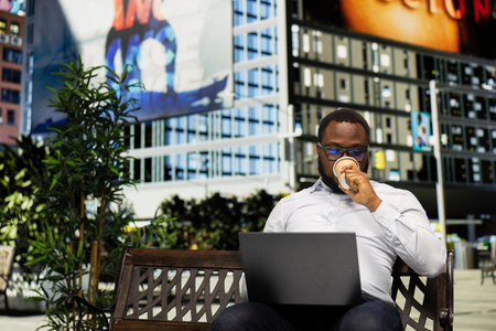 Black male adult working on laptop from bench and serving a coffee cup in the park, reading emails and reports in the downtown area. Skyscrapers near avenue, metropolitan setting.の写真素材