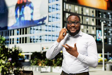 Black adult male chatting on a phone call through busy urban avenue, showcasing casual lifestyle and street style with modern architecture. Dynamic rhythm of city life around him.の写真素材