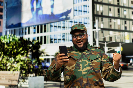 Professional smiling military man reading satisfying good news on mobile phone, cheering in the city...