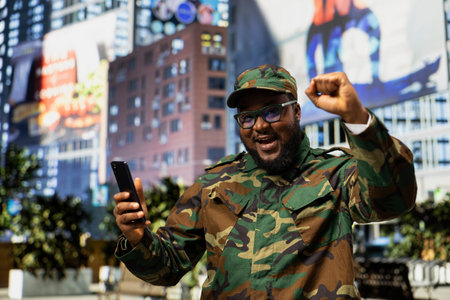 African american happy military personnel reading satisfying good news on mobile phone, cheering in the city center and feeling happy with his accomplishment. Honor, loyalty and security.の写真素材