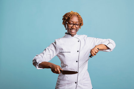 Smiling professional cook stands confidently against isolated background, holding knife and meat fork. Happy woman chef demonstrates culinary expertise, showing pride in cutting skills and cooking.の写真素材