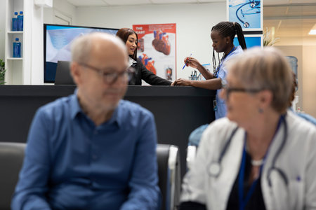 Elderly man sits with senior doctor in waiting area as asian receptionist and black nurse review...