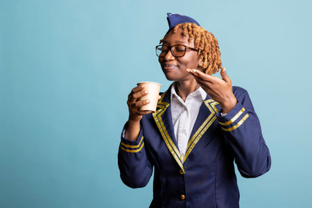 Black woman in flight attendant uniform enjoys aroma of freshly brewed coffee before takeoff. Calm airline stewardess grasps disposable cup gently, appreciating quiet moment before departure.の写真素材