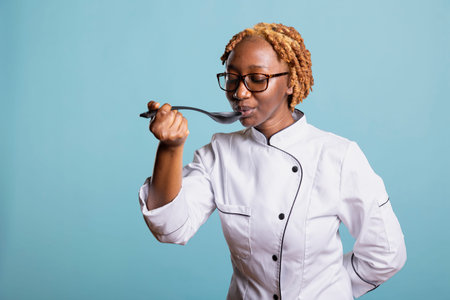 Black woman chef enjoys tasting freshly prepared dish, checking seasoning and ingredients with confidence. Young professional cook evaluating flavor and aroma from plastic cooking spoon.の写真素材