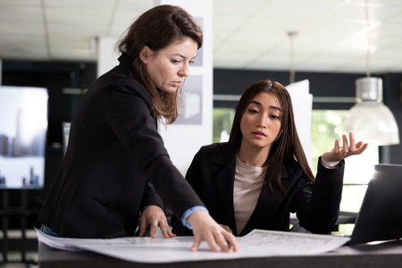 Caucasian architectural specialist points to printed design sheets on desk, discussing completion...