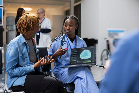 Young nurse using laptop to review digital medical records with african american woman in hospital lobby. Health specialist explains brain scan results with female patient and provides treatment plan.の写真素材