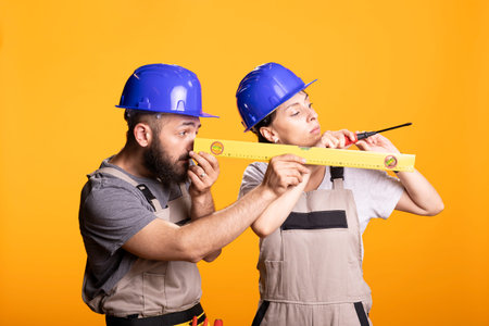 Bearded man examines yellow measuring level as woman works with screw, showing teamwork in construction industry. Male builder focuses carefully on accuracy, precision and professional craftsmanship.の写真素材