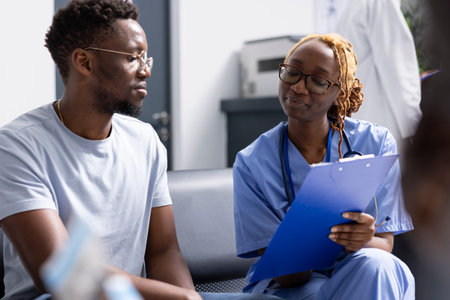Black nurse sits next to young man in hospital lobby to explain medical tests and upcoming procedures. Female healthcare worker with clipboard, providing supportive advice to encourage healthy habits.の写真素材