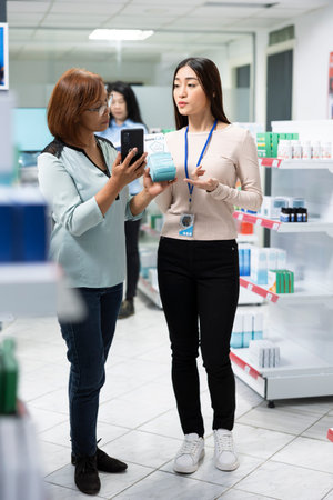 Woman shows her smartphone screen to pharmacist, standing in store aisle filled with pharmaceutical...