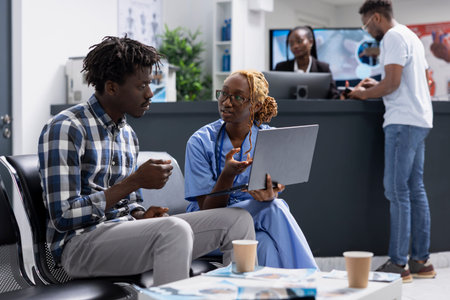 Black man patient sits with healthcare specialist in medical facility, engaged in focused conversation about test results. Nurse using laptop to assist clinic visitor with health insurance questions.の写真素材