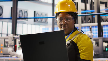 Solar panels plant technician uses laptop to oversee systems, ensuring machinery operates at optimal...