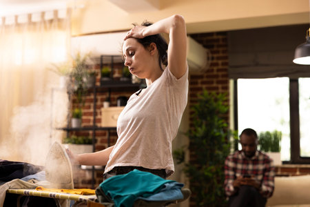Exasperated white woman holding head and iron, looking at pile of clothes left to do, feeling tired from housework. Young female homeowner showing exhaustion while completing daily chores.の写真素材