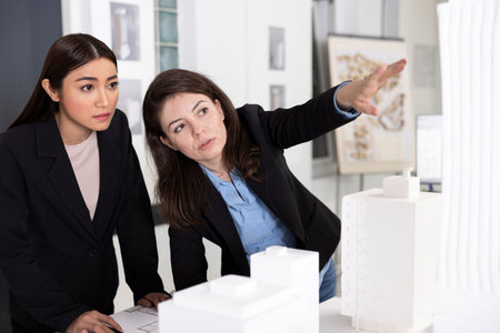 Architects analyze detailed architectural model while discussing structural ideas and project layout. Professional women show teamwork, precision and creative problem solving in modern design office.の写真素材