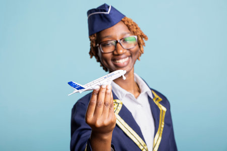 Playful flight attendant holds commercial scale plane, standing in studio with blue background. Joyful woman represents aviation dreams, global travel and passion for airline service and tourism.の写真素材