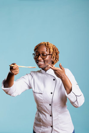 Joyful female chef in uniform tasting food using wooden spoon against blue background. Professional cook expressing satisfaction and creativity, showing passion and excellence for culinary arts.の写真素材