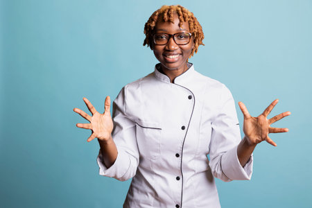 Smiling female chef in uniform gestures with open hands while standing in blue background studio. Professional cook expresses friendliness and enthusiasm, creating welcoming and positive culinary vibeの写真素材