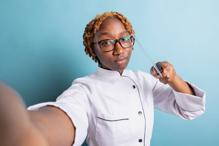 African American professional cook playfully pointing kitchen knife toward camera, recording culinary video. Confident female chef shows seriousness in preparation for gourmet challenge.の写真素材