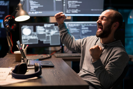 Joyful bearded programmer raising fists in celebration after solving debugging issue in tech workspace. Excited male software developer achieving coding breakthrough during night shift.の写真素材