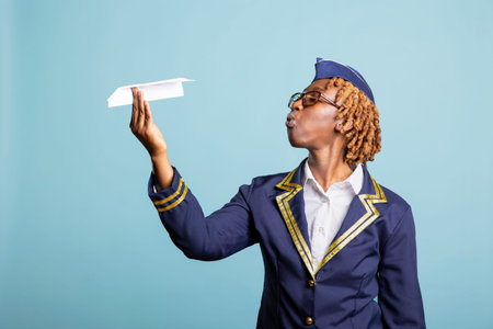 Black airline stewardess with glasses blows air at diy aircraft, enjoying fun and lighthearted moment against isolated background. Playful air hostess in uniform playing with paper airplane in studio.の写真素材