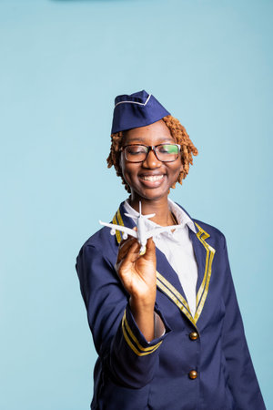 Happy female cabin crew member wearing elegant airline uniform and holding model aircraft in studio. Cheerful black woman with toy airplane promoting travel deals and aviation career.の写真素材