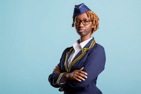 Elegant flight attendant in navy uniform stands with arms crossed against blue background. Air hostess with glasses looking at camera, expressing confidence, authority and pride in aviation industry.の写真素材