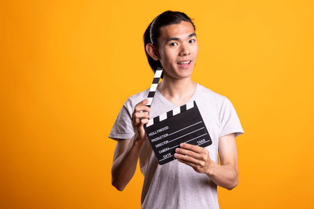 Smiling Asian man dressed casually stands in studio holding film slate and looking at camera....