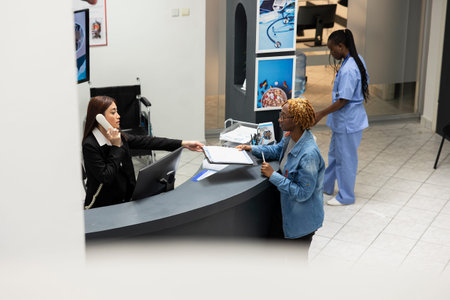 Female clinic administrator multitasks, having phone call and attending to african american patient at front desk. African American woman receiving registration forms on clipboard from receptionist.の写真素材